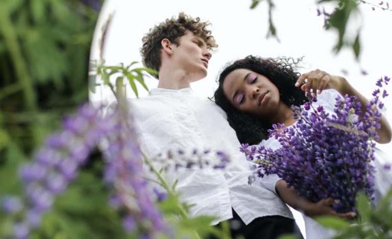 A round mirror set in a field of flowers reflects a serene white man and black woman dressed in white holding purple flowers