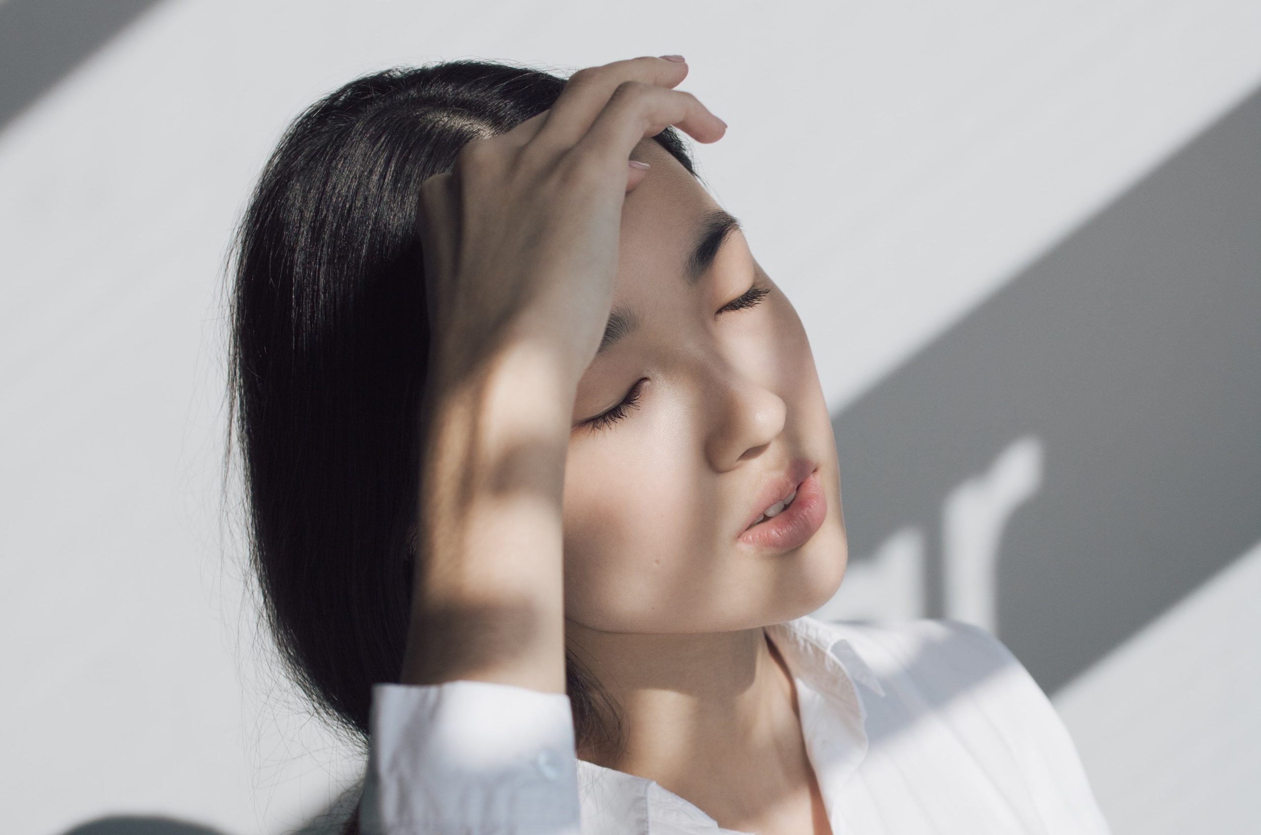 A Korean girl in a white shirt with her eyes closed and her hand by her forehead