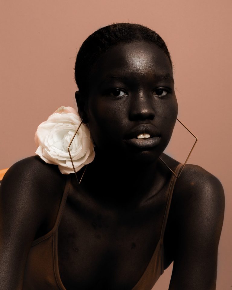 A striking black model wearing diamond wire gold hoop earrings, with a large white rose on her right shoulder